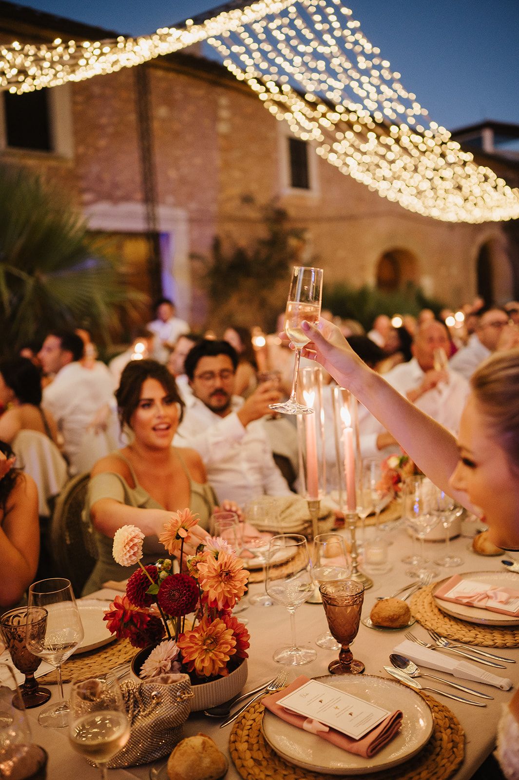 Wedding guests raise champagne glasses under fairy lights at elegant outdoor dinner reception.