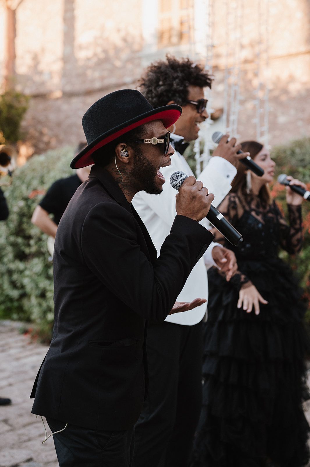 Energetic wedding band performer in black suit and wide-brim hat sings into microphone at outdoor reception.