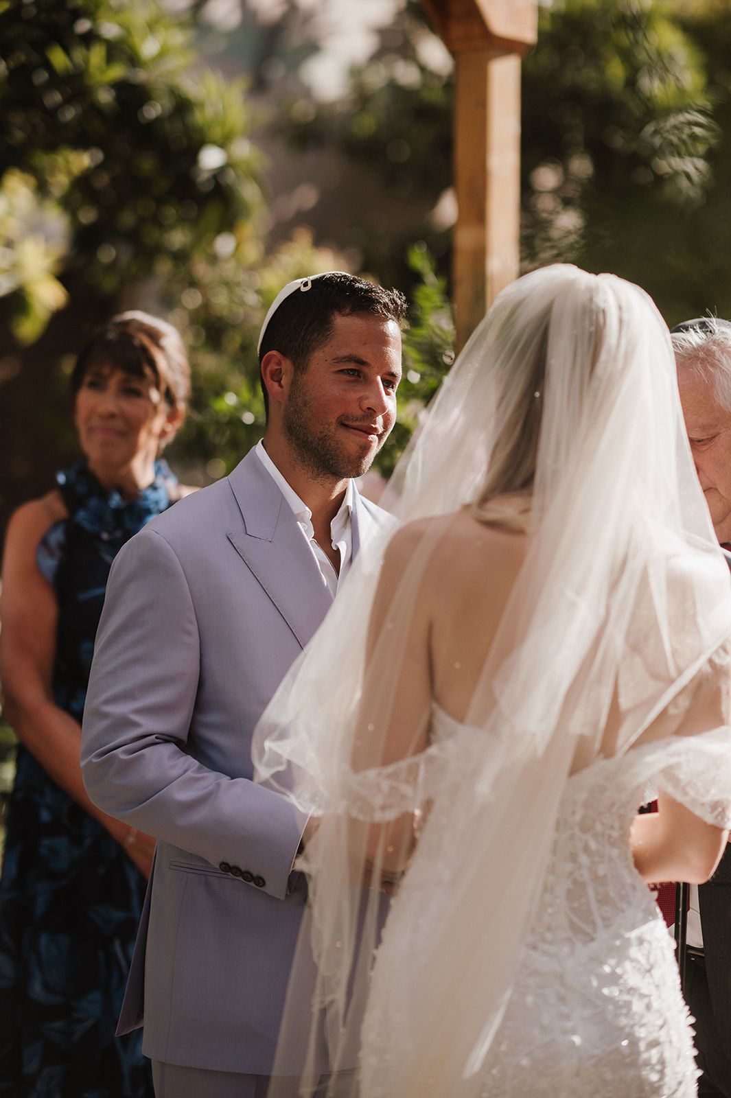 Groom in a light gray suit smiles at his bride in a lace wedding dress and veil during their outdoor ceremony.
