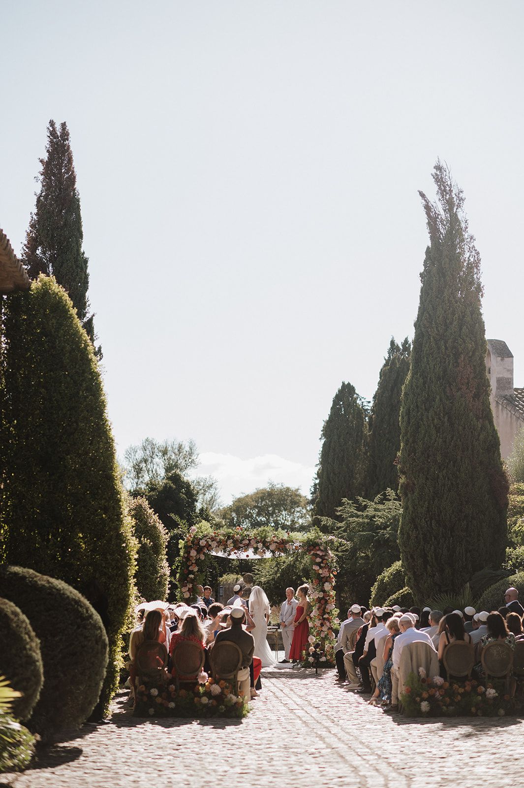 Guests seated in rattan chairs along a cobblestone aisle surrounded by cypress trees during an outdoor garden wedding ceremony.