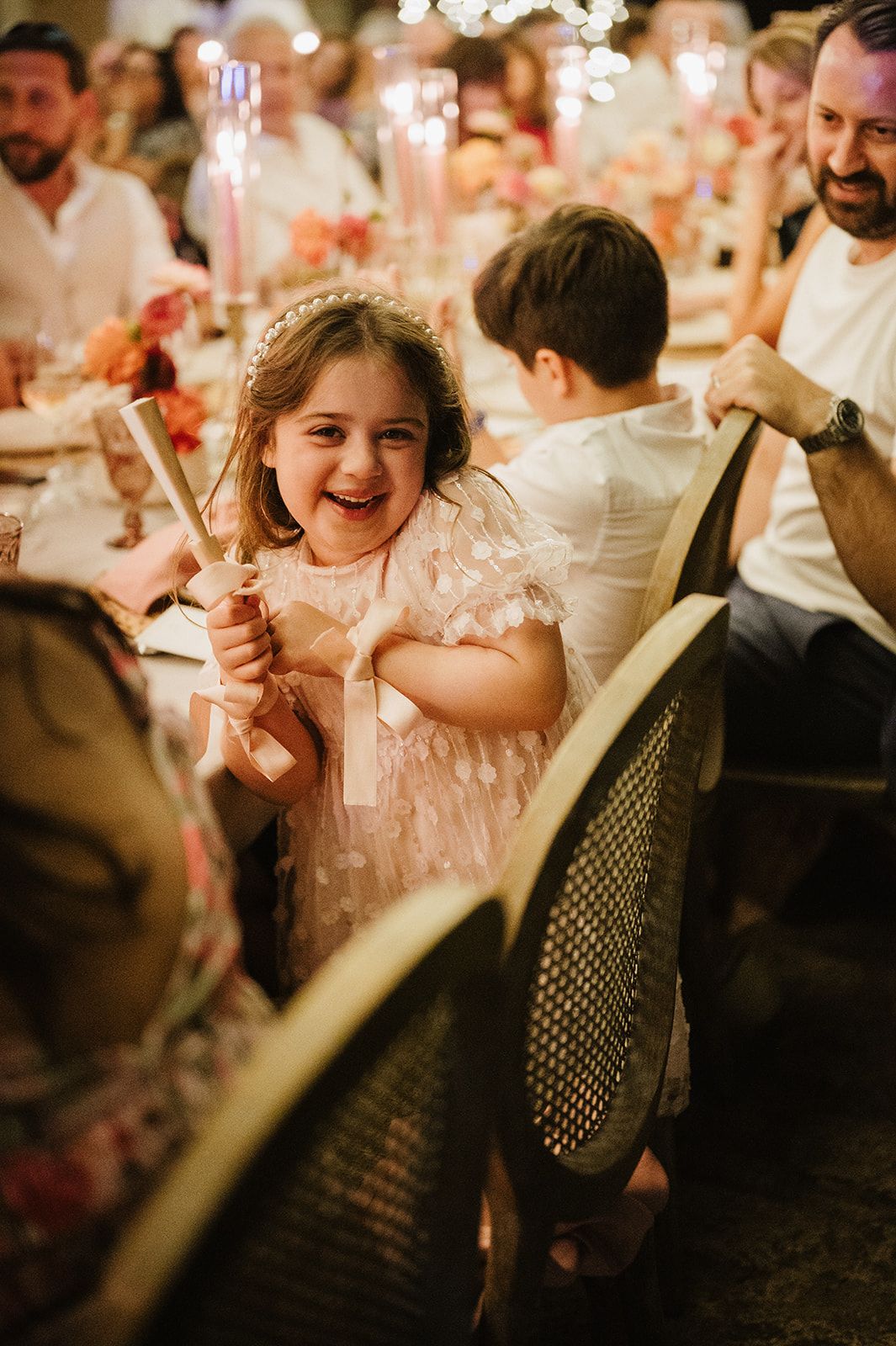 Adorable flower girl in pink dress laughs while holding a party popper at elegant candlelit wedding reception.