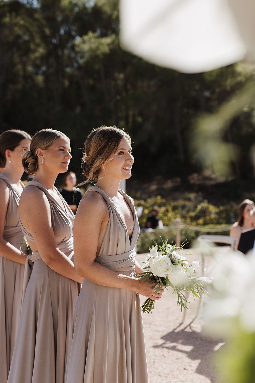 Smiling bridesmaids in taupe multiway dresses holding white bouquets at an outdoor wedding ceremony.