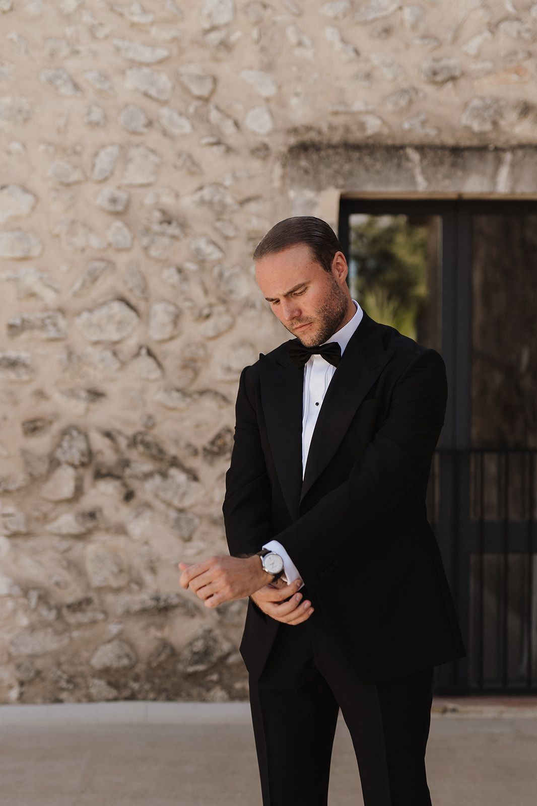Groom in black tuxedo and bow tie adjusting cufflinks outside stone building.