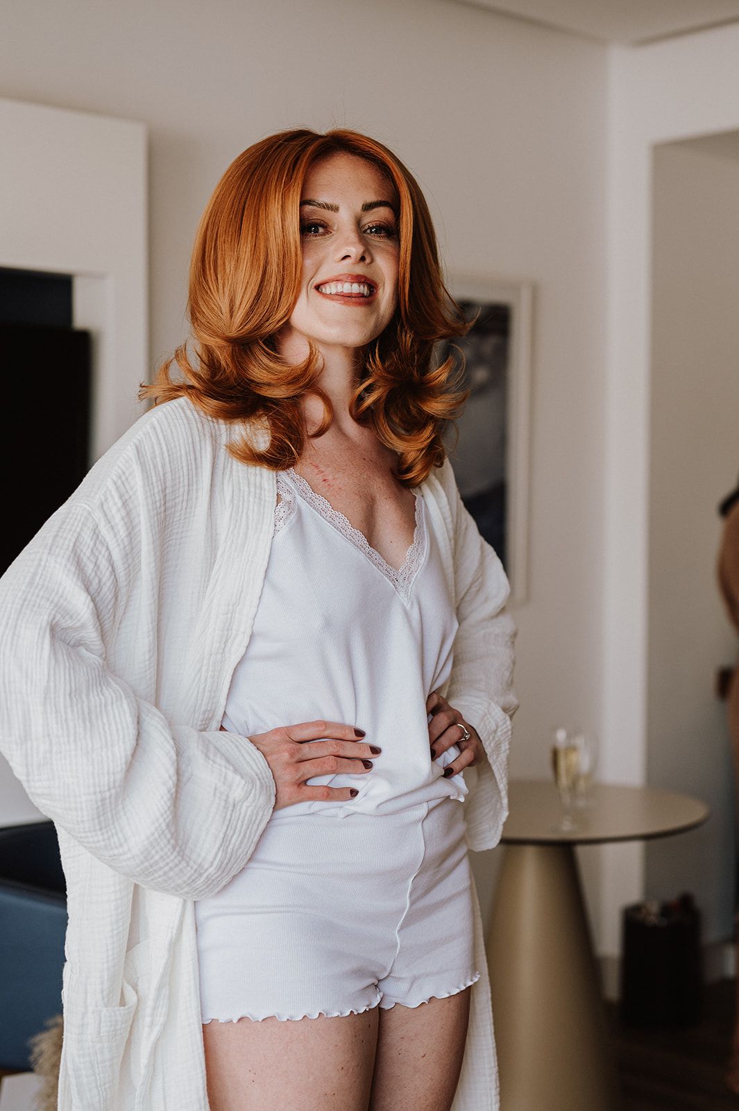 Smiling red-haired bride in white silk shorts and open robe posing in bright hotel room.