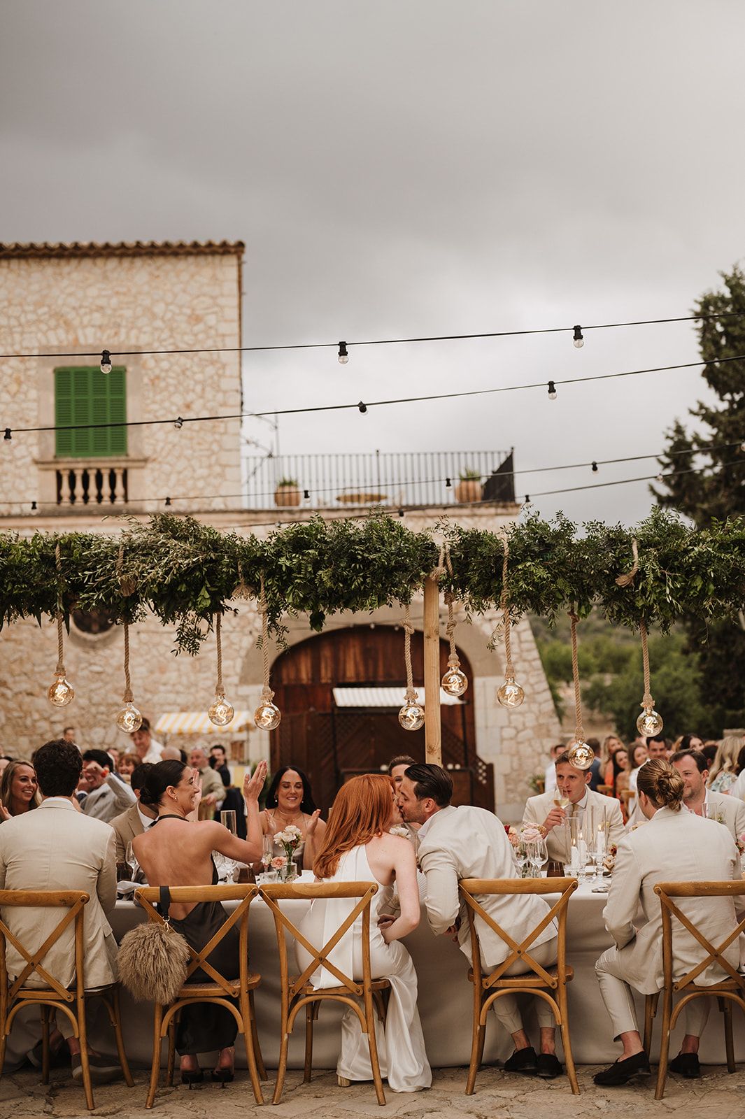 Wedding guests celebrate at a rustic stone venue with hanging globe lights, greenery garlands, and wooden cross-back chairs.
