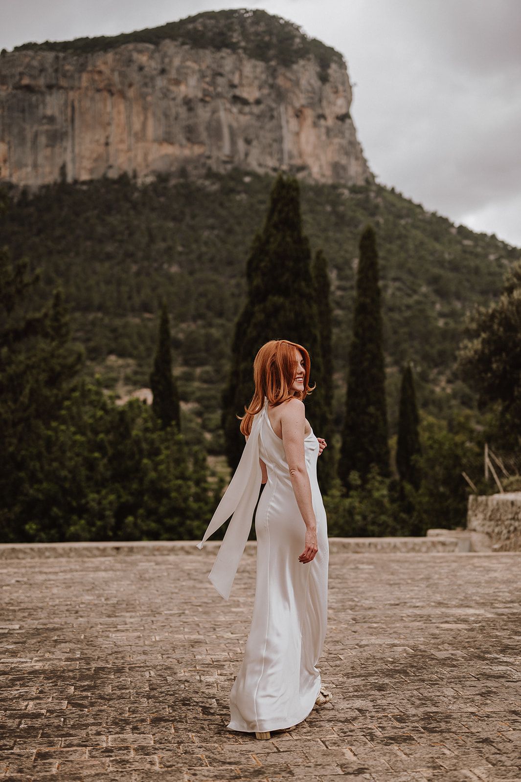 Redhead bride in elegant white halter gown poses on stone terrace with mountain scenery behind her.