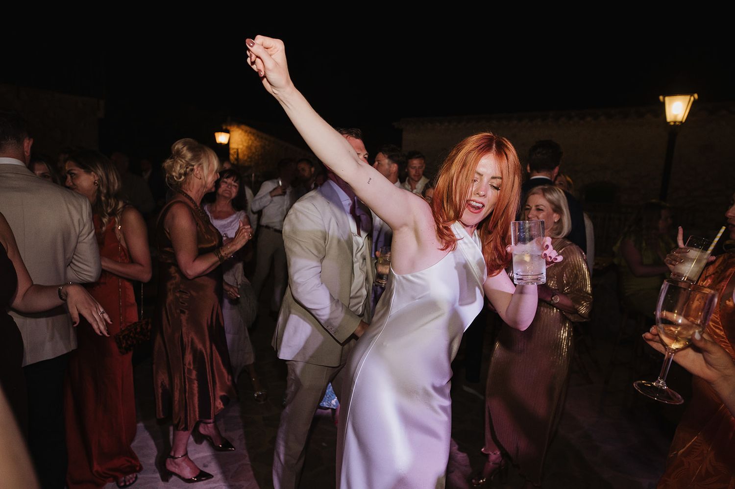 Red-haired woman in white satin dress dancing joyfully at an outdoor wedding reception.