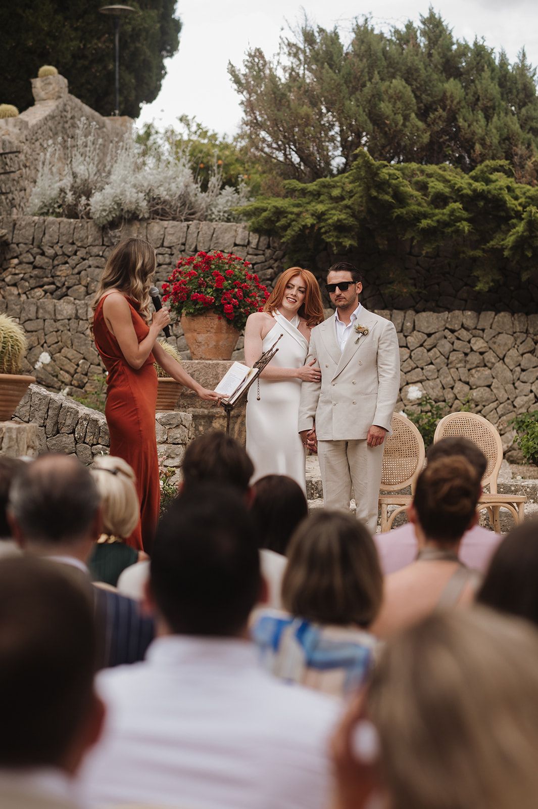 Bride in white dress and groom in beige suit stand with officiant during outdoor wedding ceremony.