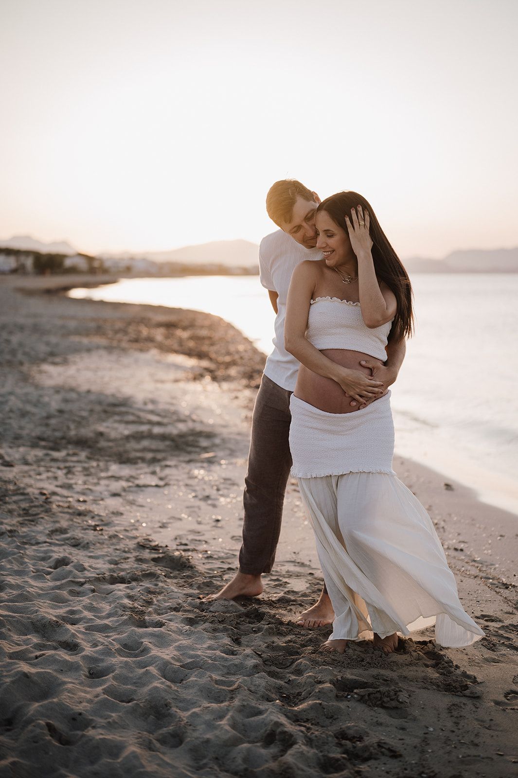 Couple embracing on sandy beach at sunset, pregnant woman in white dress with partner holding her belly.