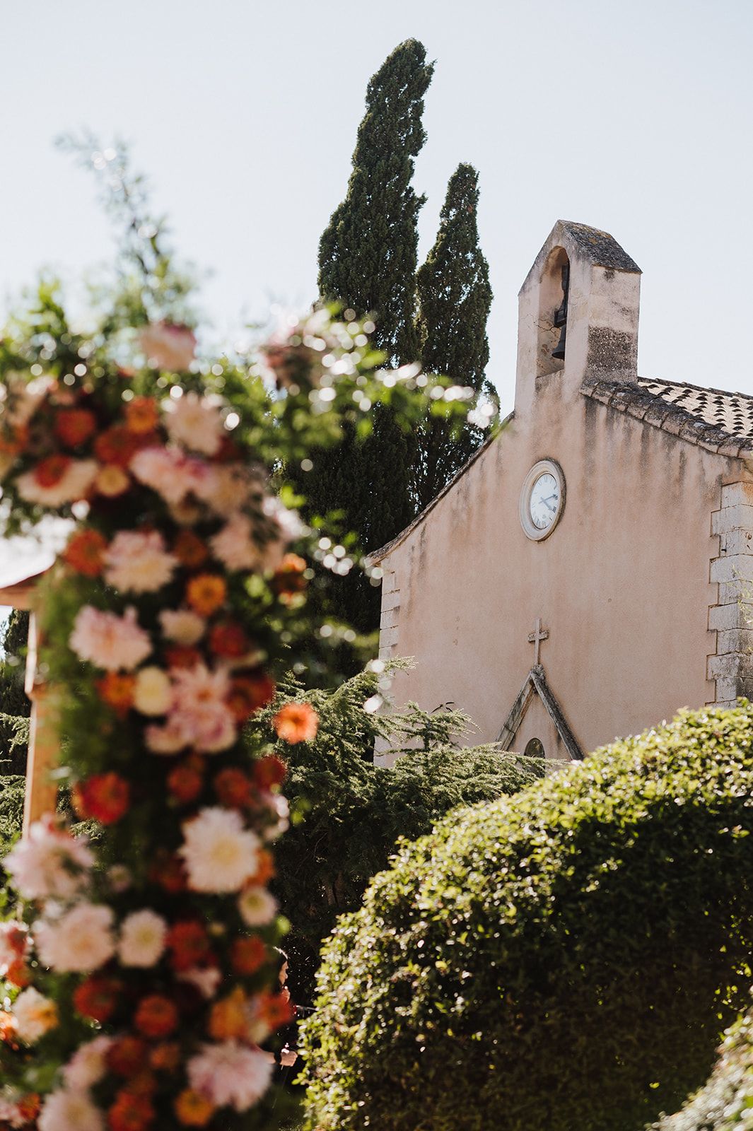 Colorful floral arch with pink and orange blooms frames rustic church with clock tower at wedding.