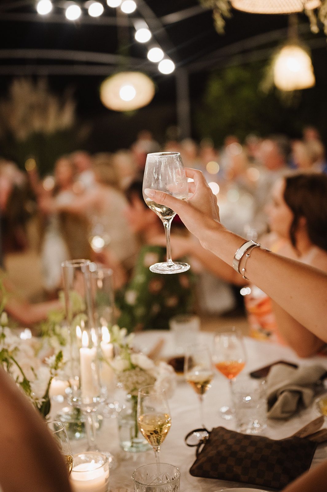 Guest raises wine glass in toast at romantic candlelit outdoor wedding reception dinner celebration.