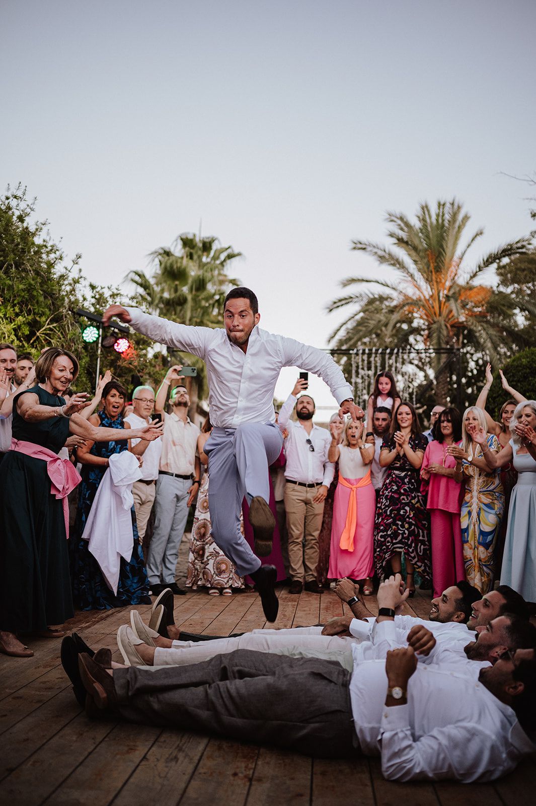 Groom jumps over guests lying on dance floor at lively outdoor wedding reception.