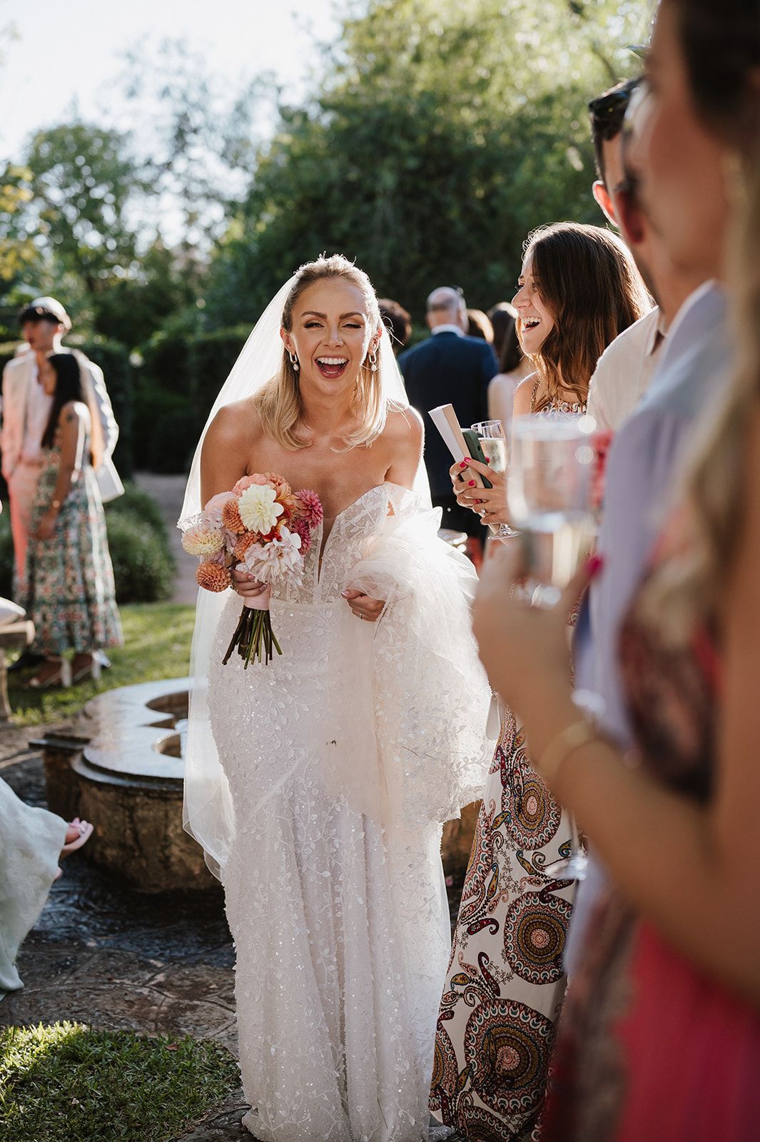 Radiant bride holds bouquet laughing with guests at outdoor reception.