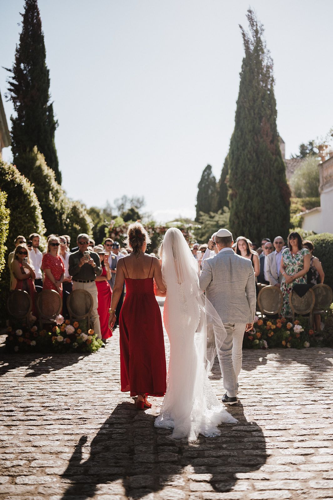 Bride walks down outdoor aisle with escorts at sunny garden wedding ceremony.