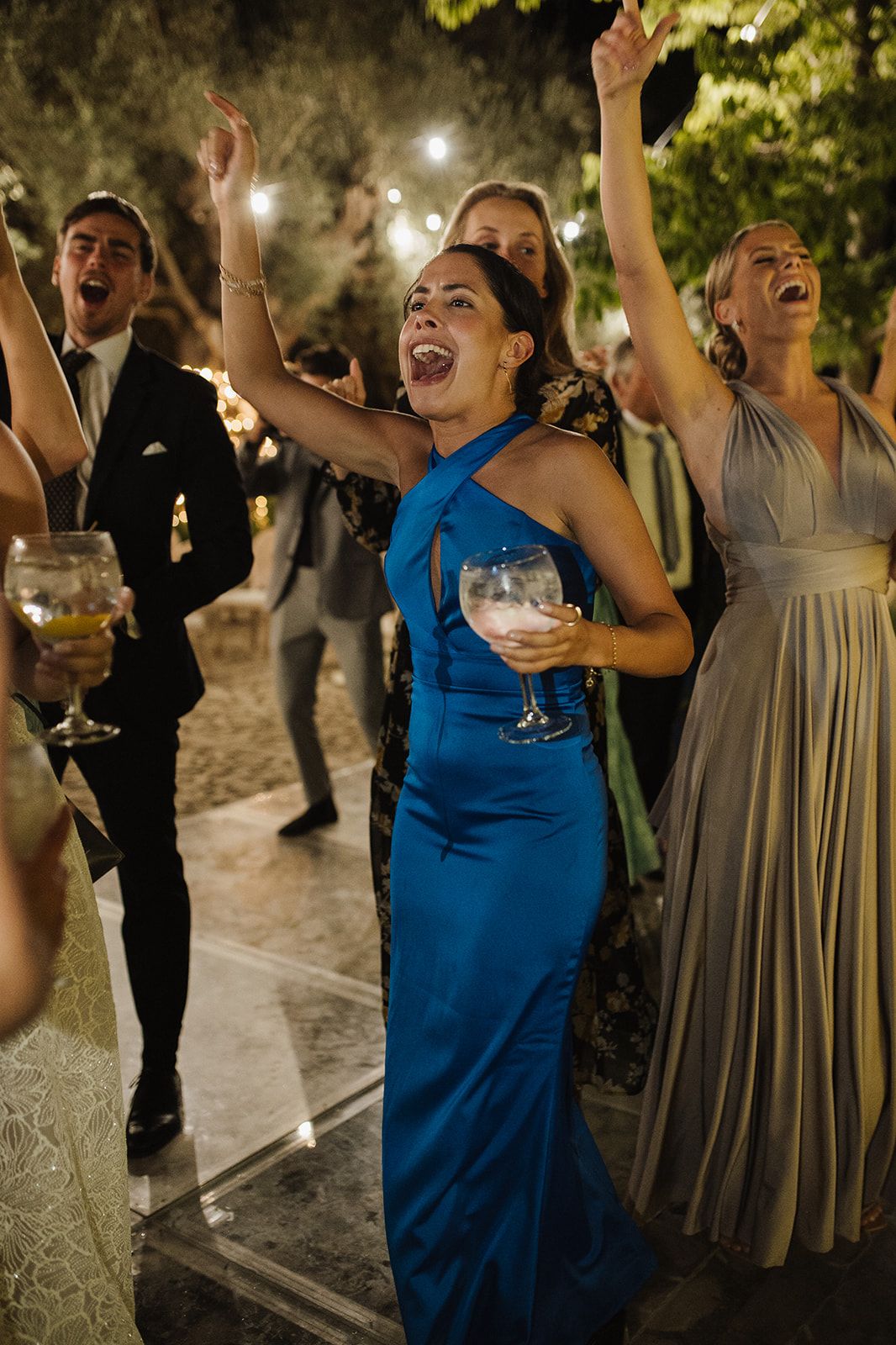 Wedding guest in blue gown dancing enthusiastically with raised arms at evening reception.
