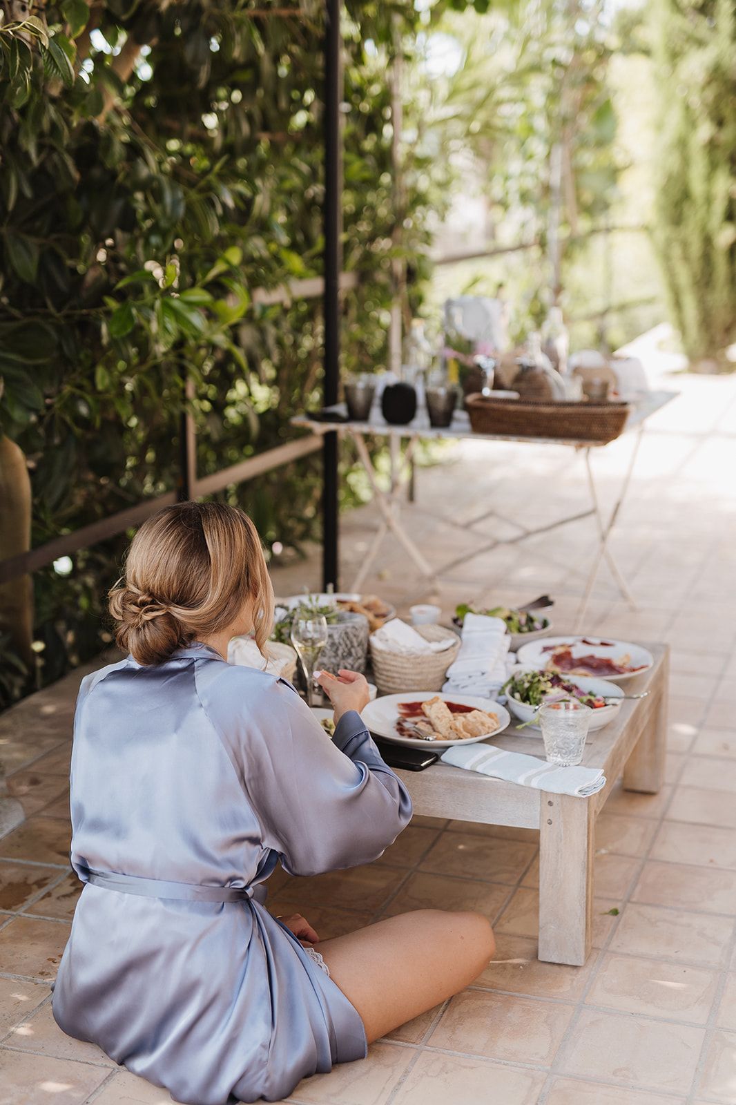Bride in silver silk robe sits cross-legged on patio floor enjoying a bridal brunch spread on a low wooden table outdoors.