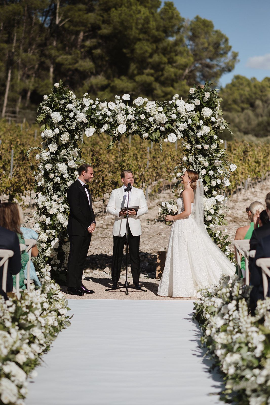 Bride and groom exchange vows under floral arch at outdoor vineyard wedding ceremony.