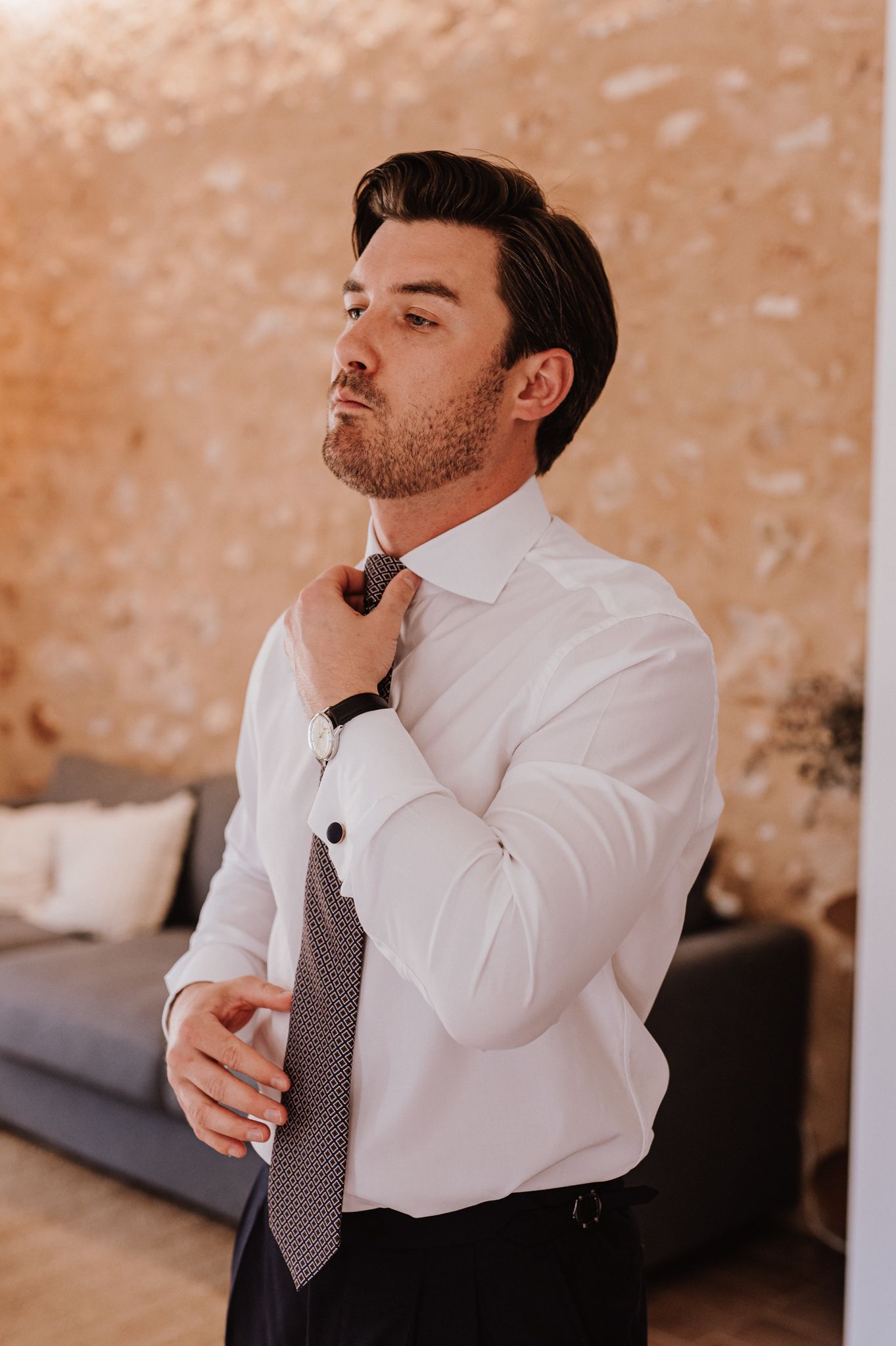 Groom in white shirt adjusting tie before wedding ceremony indoors.