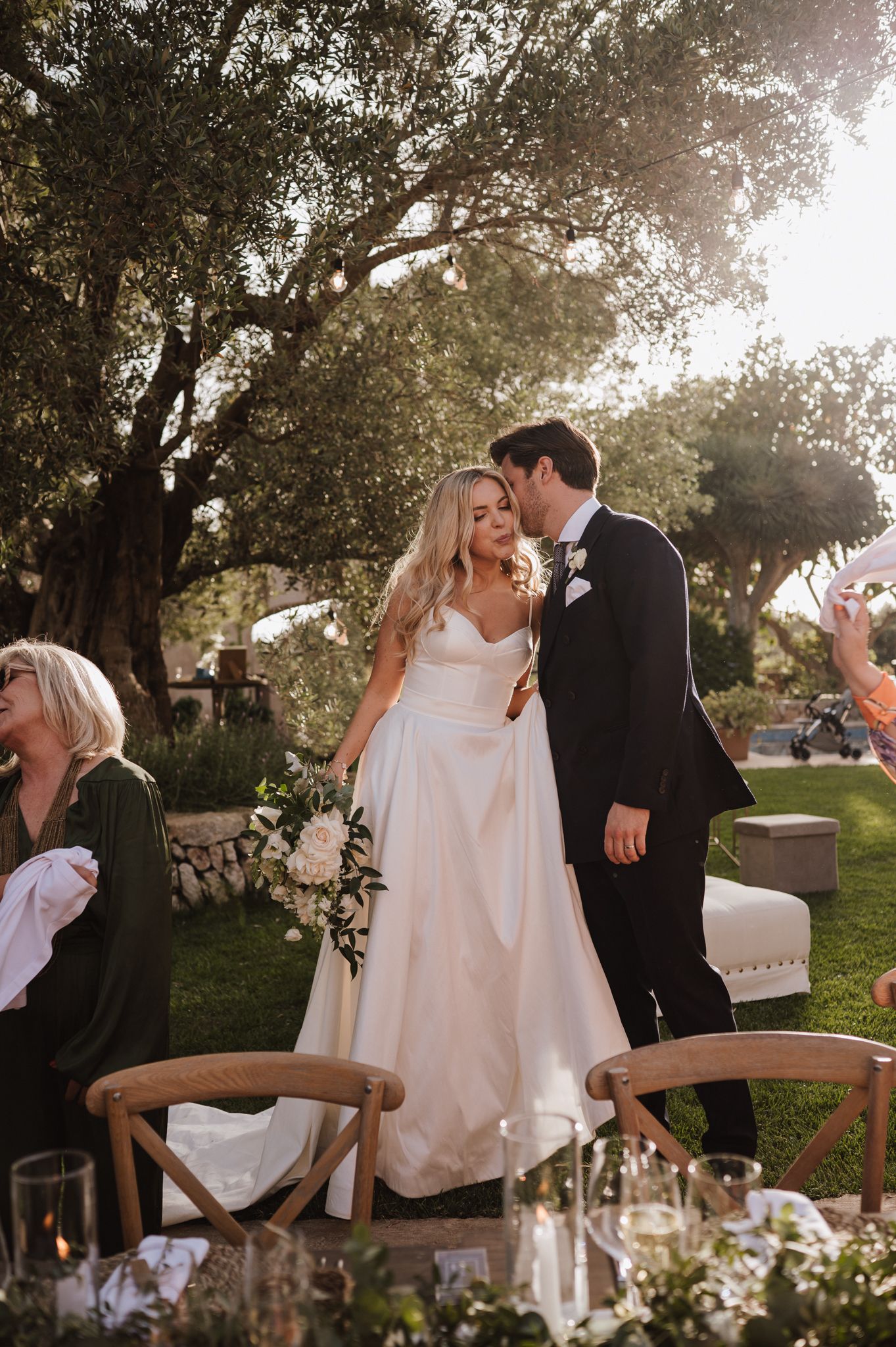 Groom kisses bride on the cheek at golden hour during an outdoor wedding reception under olive trees.