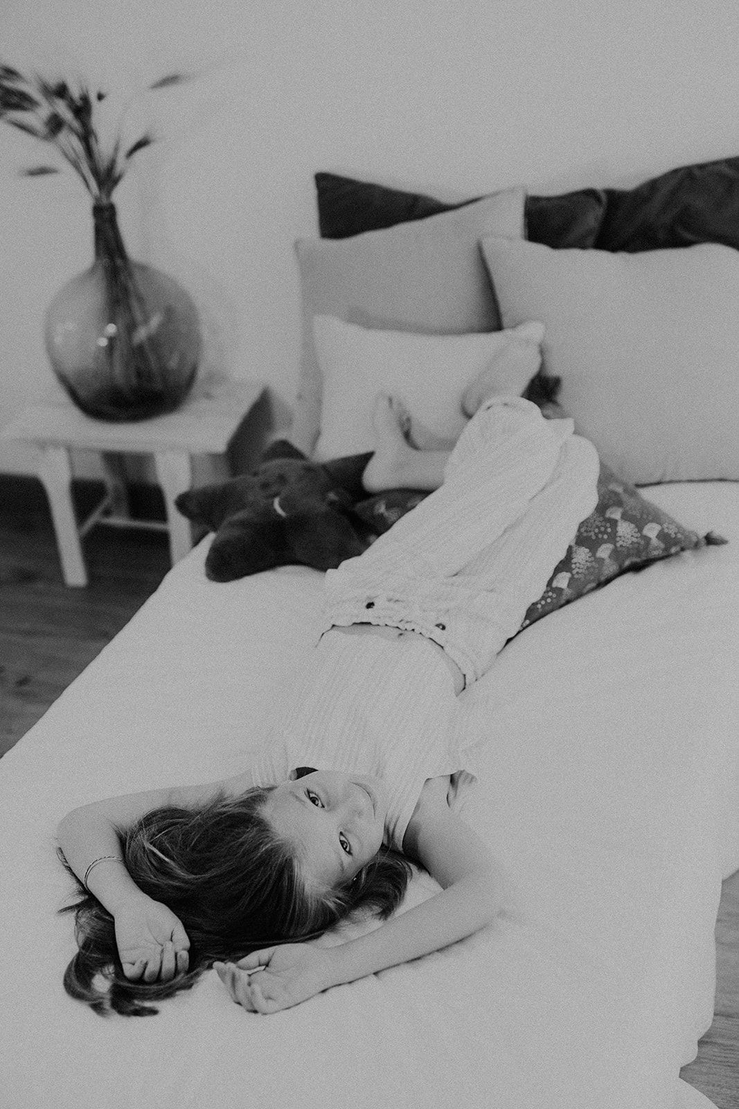 Young girl lying on white bed with cat and pillows in black and white lifestyle photo.