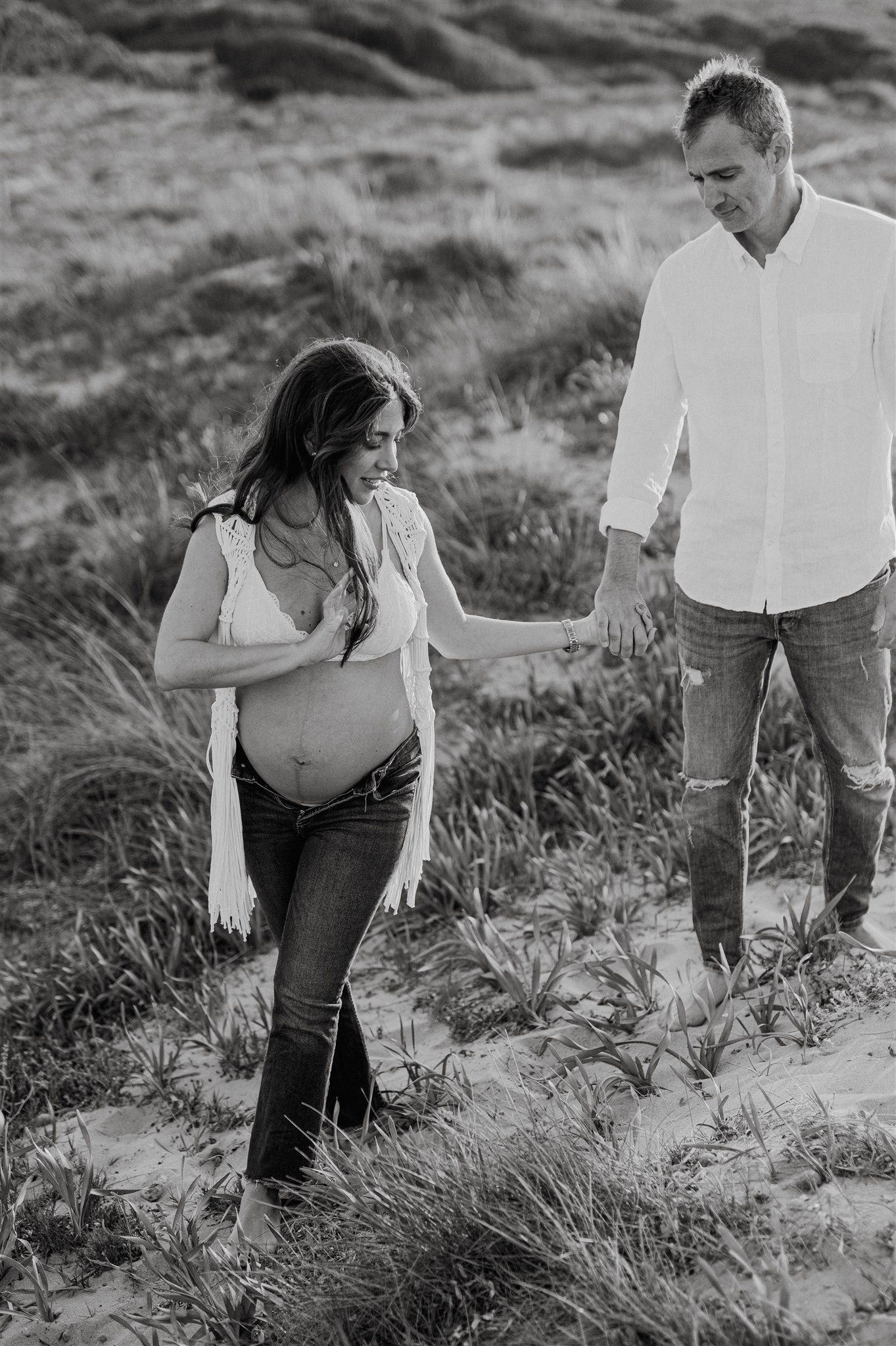 Black and white photo of a pregnant couple holding hands while walking barefoot through sandy dunes.