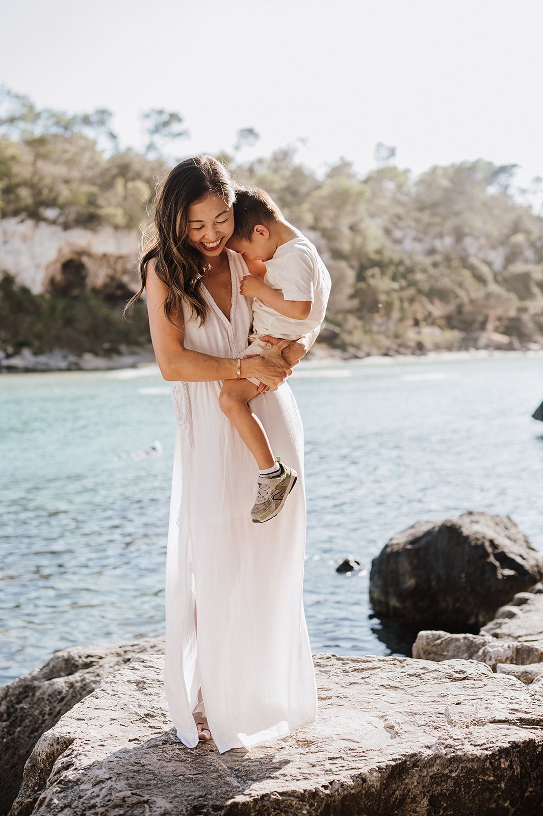A smiling woman in a white maxi dress holds a toddler on rocky shore with turquoise water behind them.