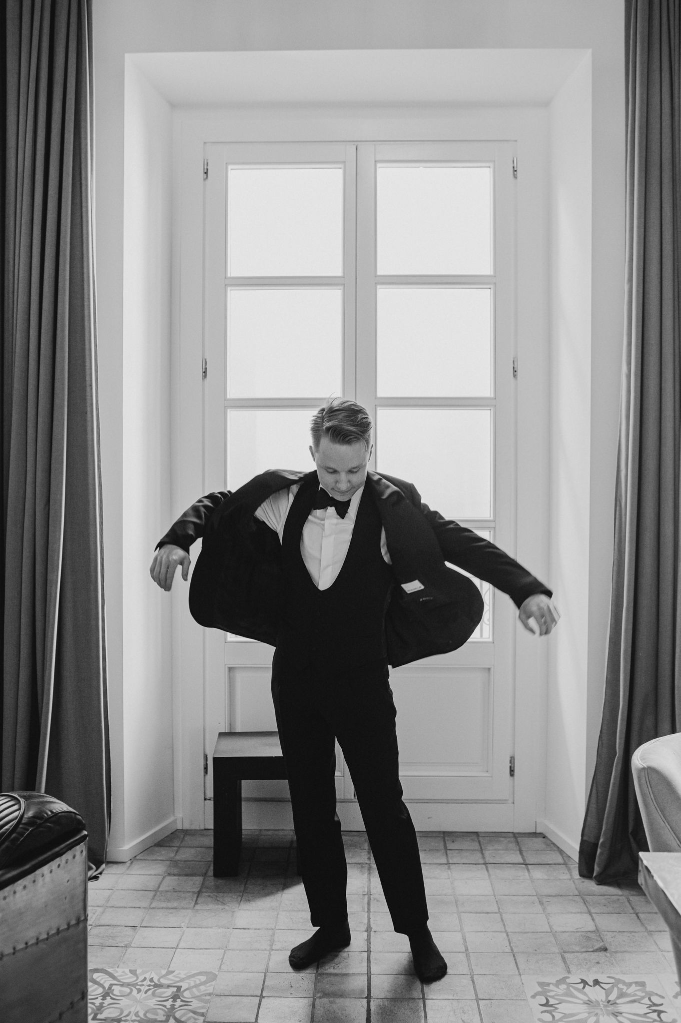 Groom in tuxedo adjusting his jacket in a bright room before wedding ceremony.