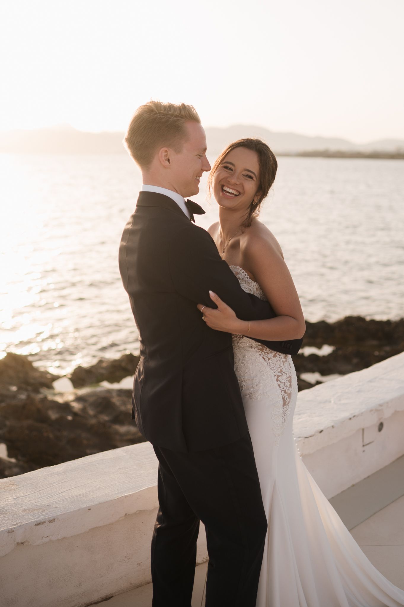 Radiant bride in fitted lace gown laughs with tuxedo-clad groom during golden hour seaside portraits.