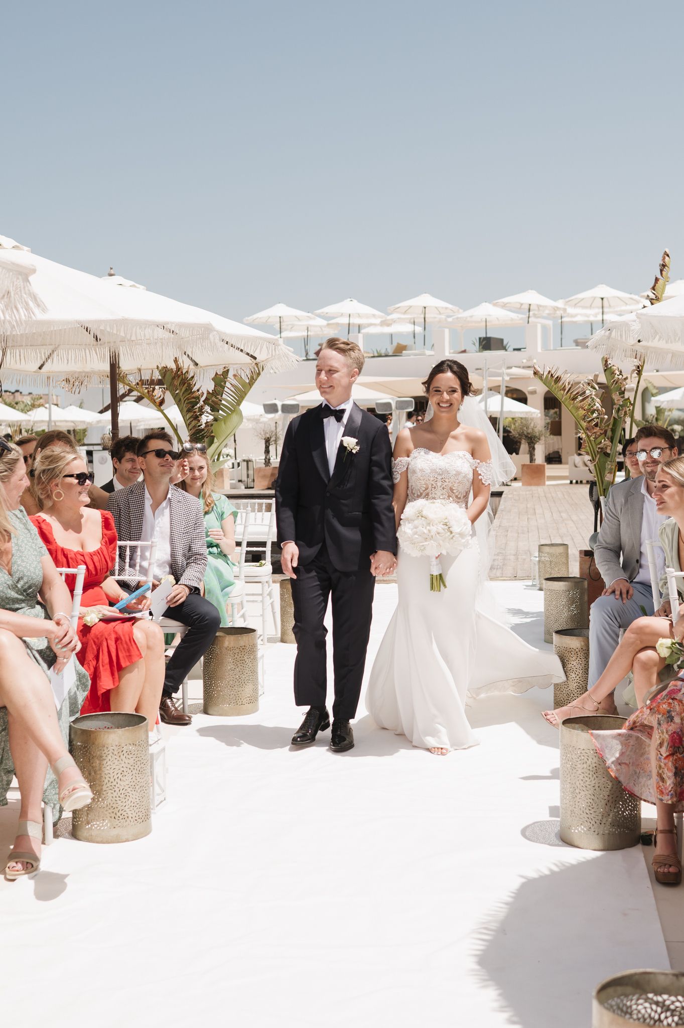 Bride and groom walk down white aisle at outdoor beach wedding ceremony with guests seated beside them.