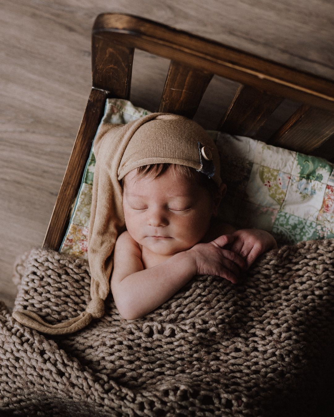 Sleeping newborn baby wearing a knit beige bonnet, wrapped in a chunky knit blanket inside a small wooden bed.