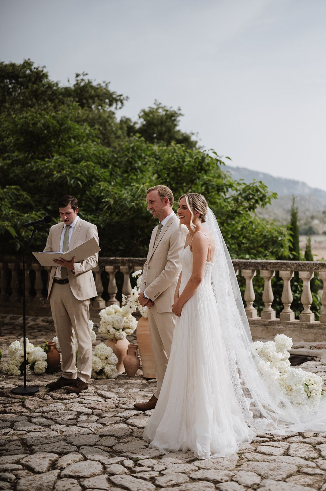 Bride and groom exchange vows on stone terrace with white floral arrangements and mountain backdrop.