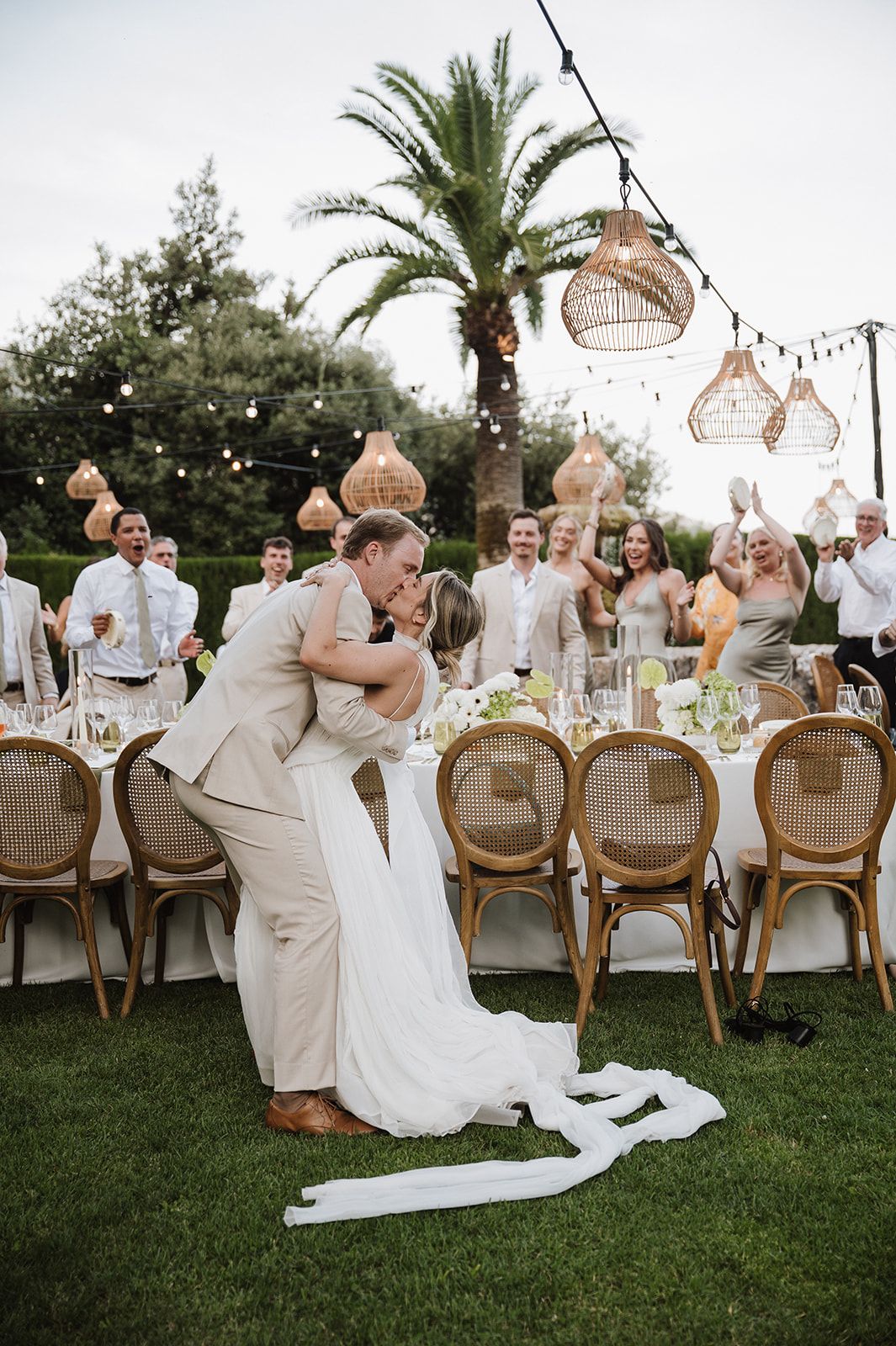 Groom dips bride for a kiss at outdoor wedding reception with guests cheering and string lights.