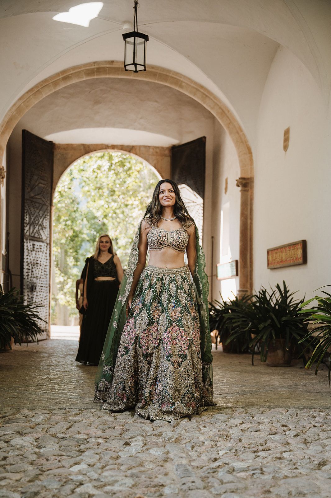Bride in stunning embroidered green lehenga walking through arched doorway at wedding venue.