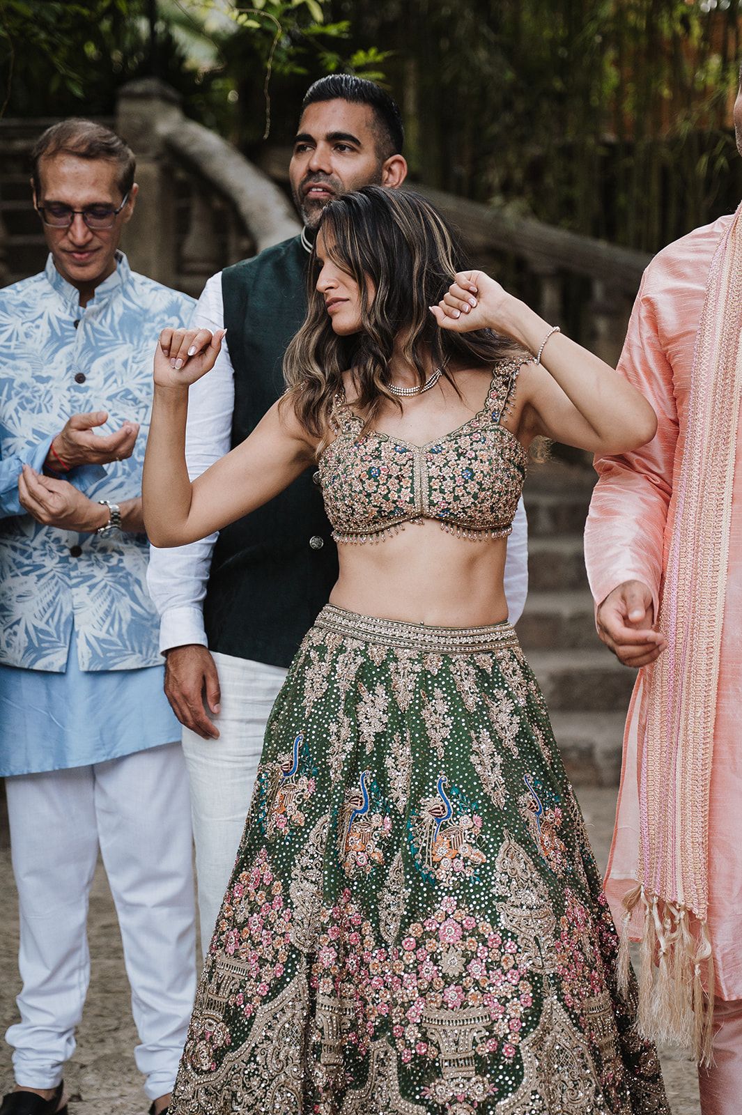 Bride dancing joyfully in embroidered green floral lehenga at outdoor Indian wedding celebration.