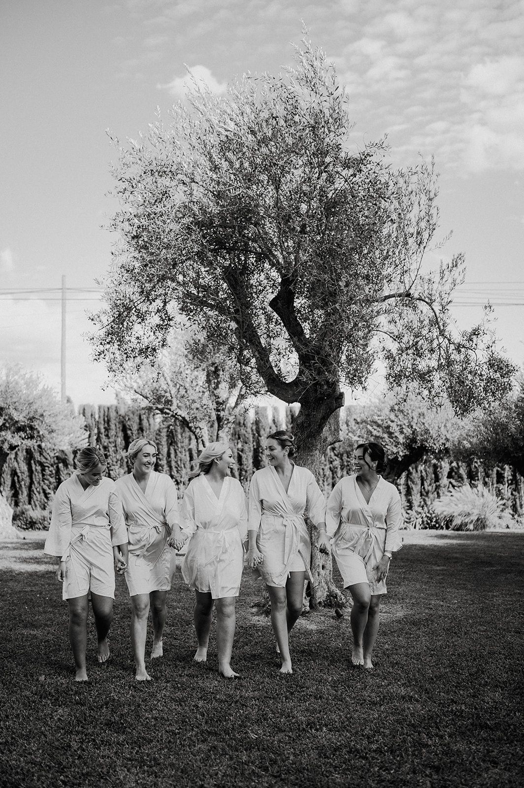 Bridal party in matching white robes walking barefoot through a lush garden with an olive tree in black and white.