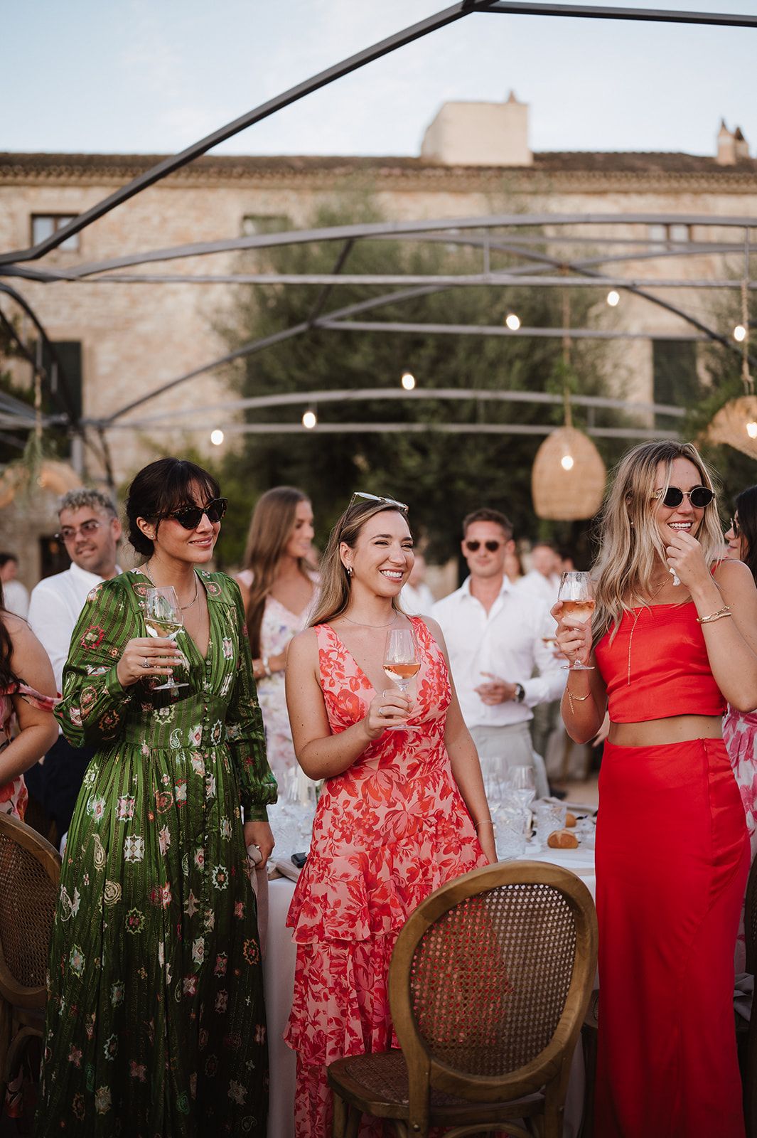 Three women in colorful floral dresses smile and hold drinks at an elegant outdoor wedding reception.