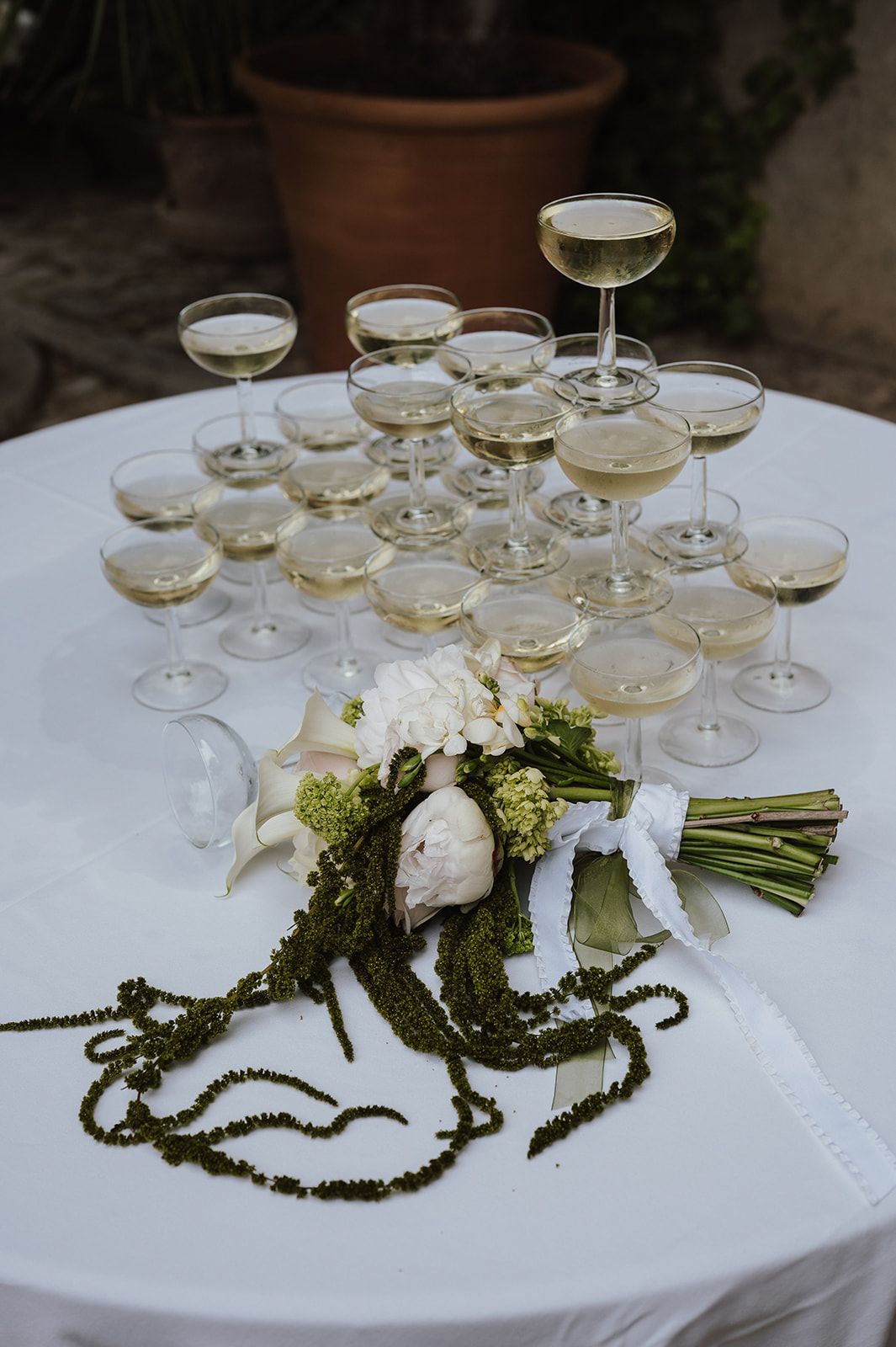 Elegant champagne tower with coupe glasses beside white floral bridal bouquet on white linen table outdoors.