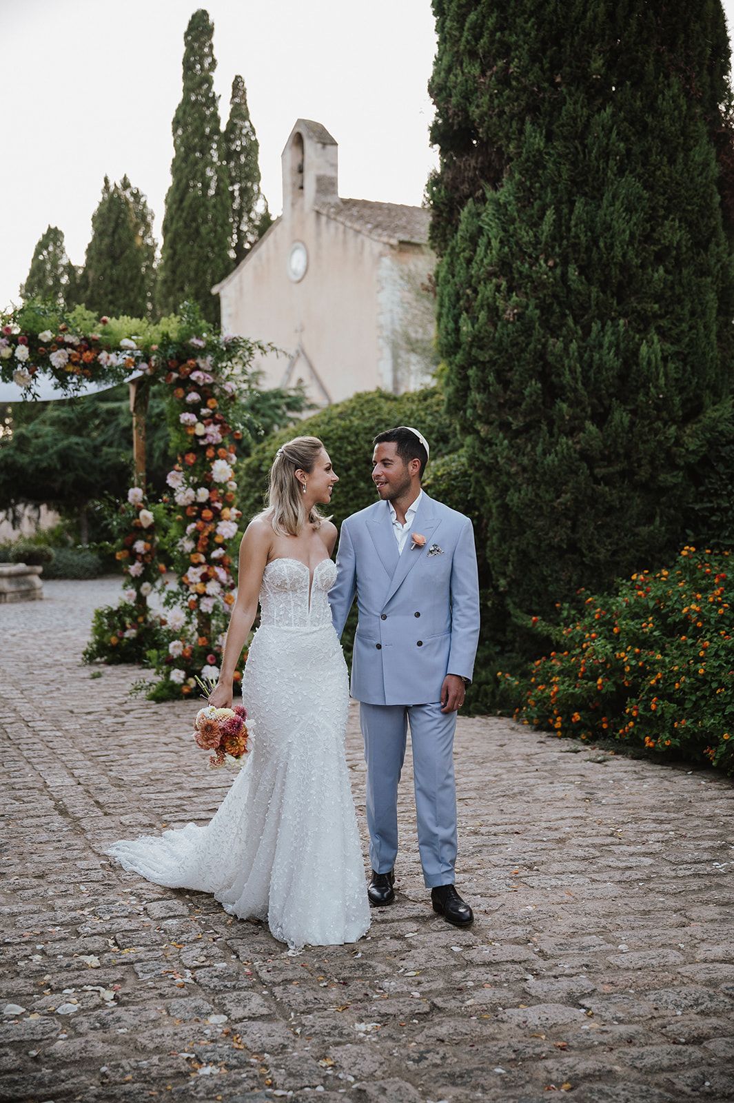 Bride and groom walk cobblestone path near floral arch at dusk.