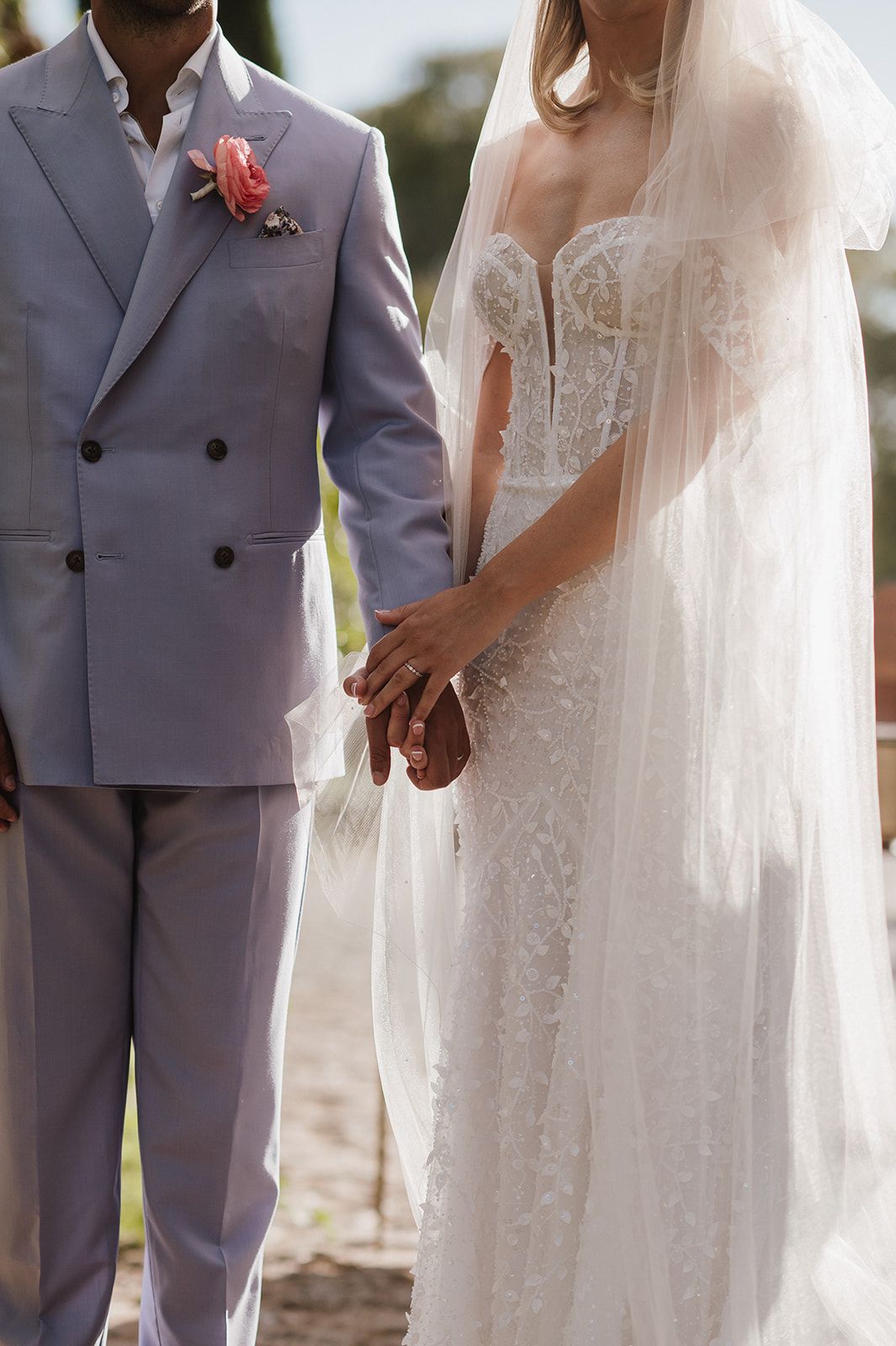 Bride in a lace off-shoulder gown and groom in a lavender double-breasted suit hold hands during their wedding ceremony.