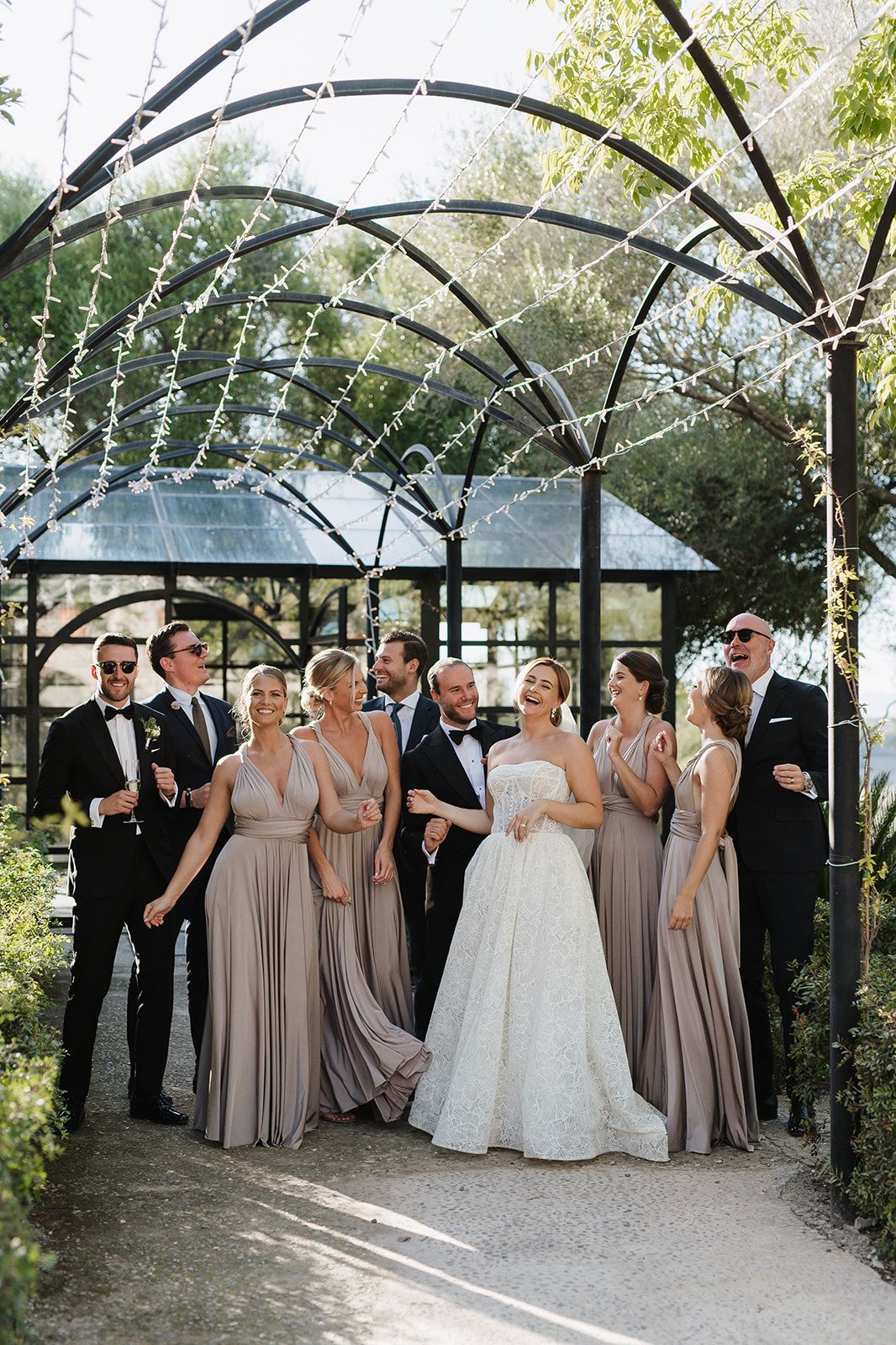 Wedding party in taupe and black attire laughs together under a decorative garden arch.