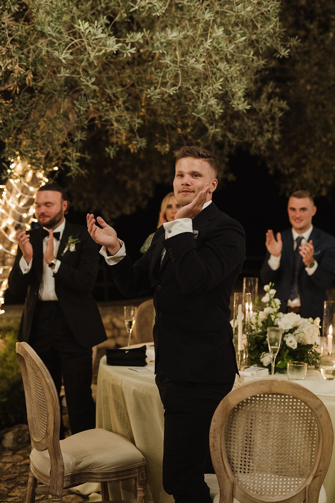Groom in black tuxedo clapping at outdoor wedding reception dinner under olive trees with string lights at night.