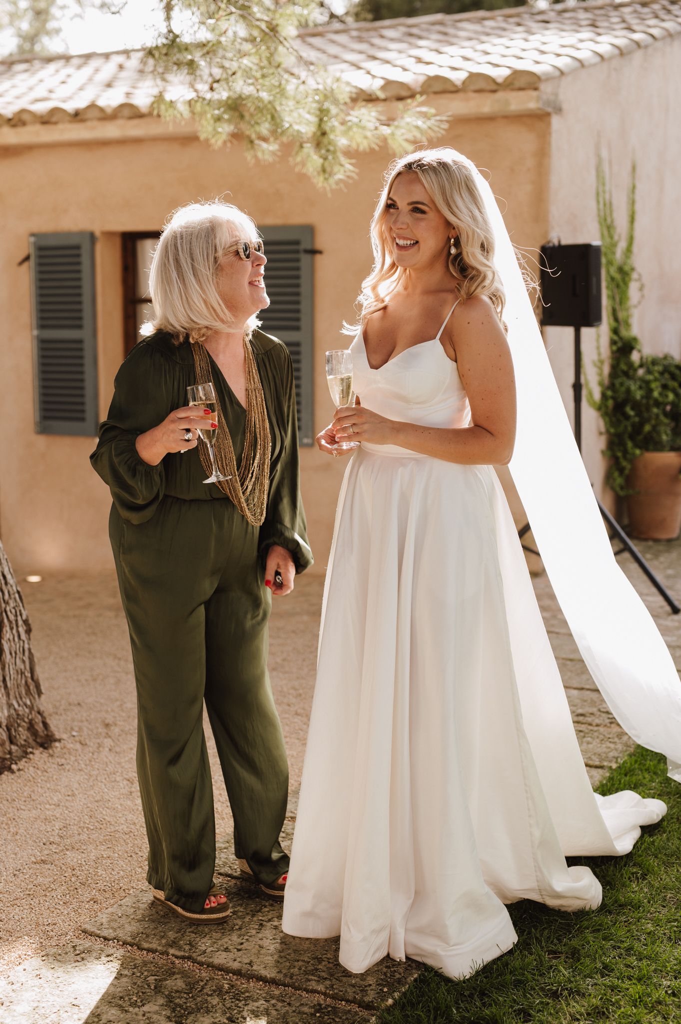 Bride in white spaghetti strap gown holding champagne with an older woman in green jumpsuit.