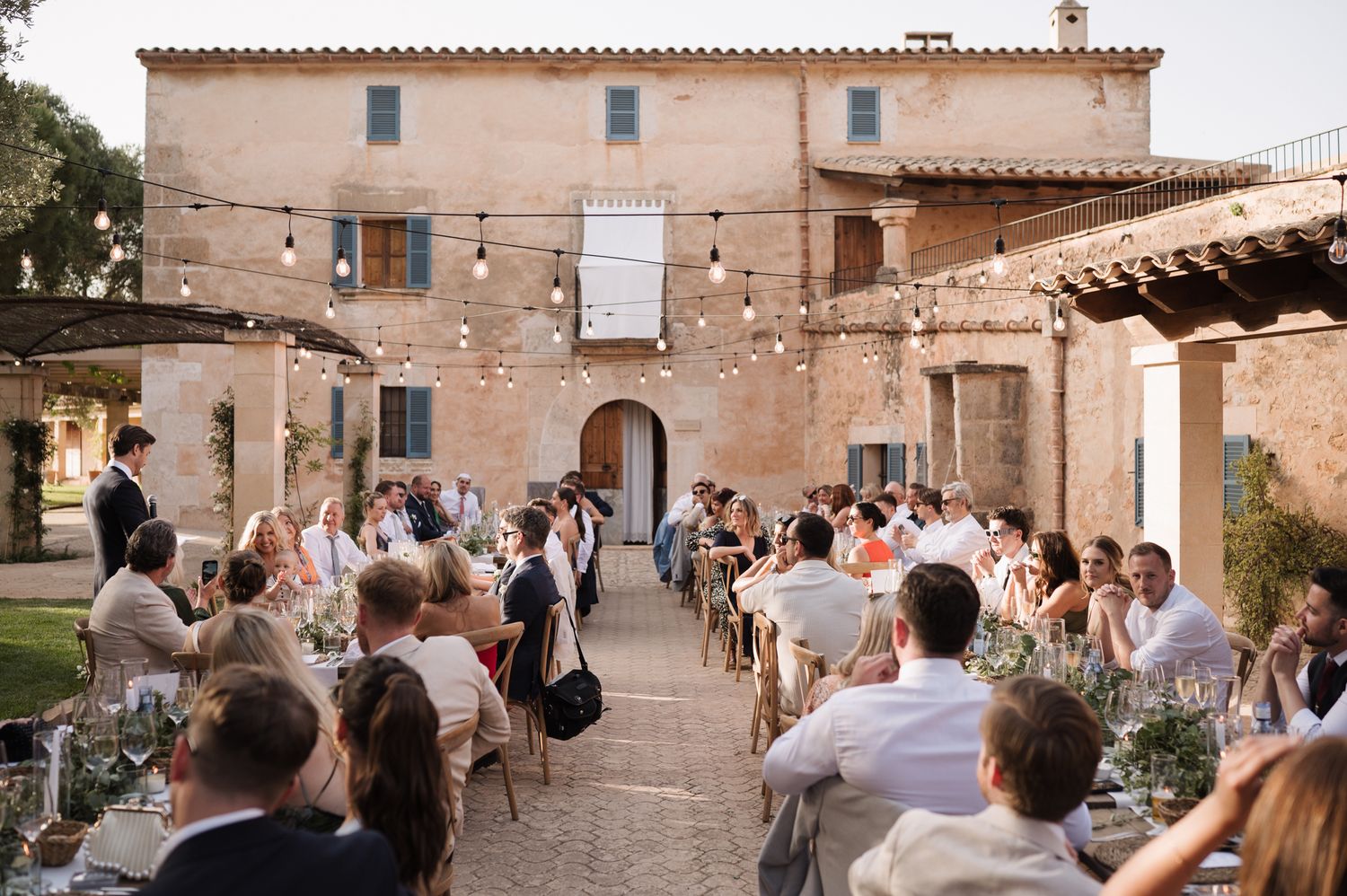Guests dining at long tables in a rustic courtyard with string lights at a Mediterranean wedding venue.