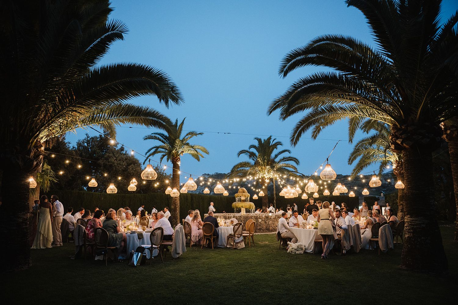 Romantic outdoor wedding reception under palm trees with warm string lights and lanterns at dusk.