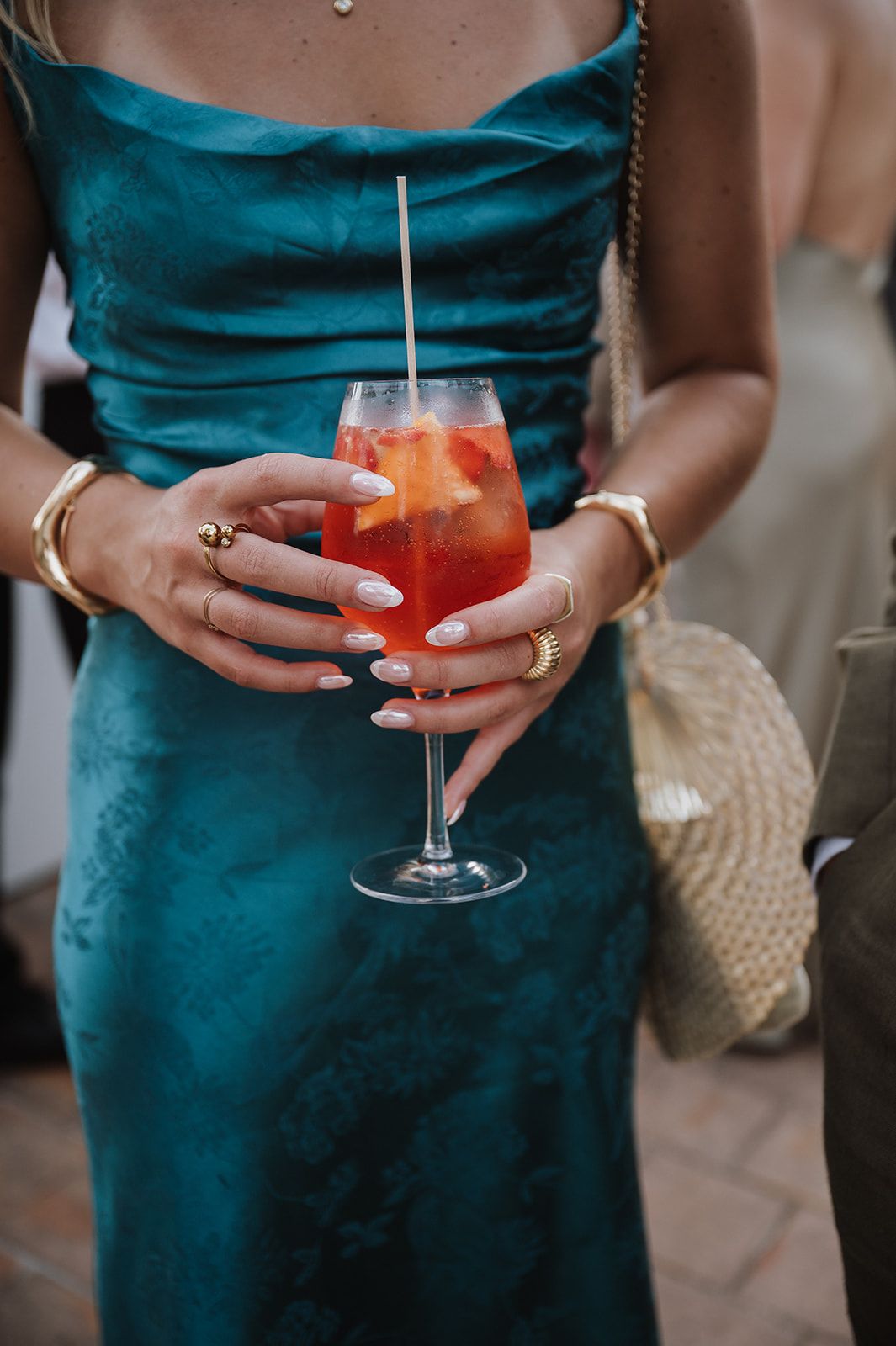 Woman in teal dress holding an Aperol Spritz cocktail with gold jewelry and white manicured nails.