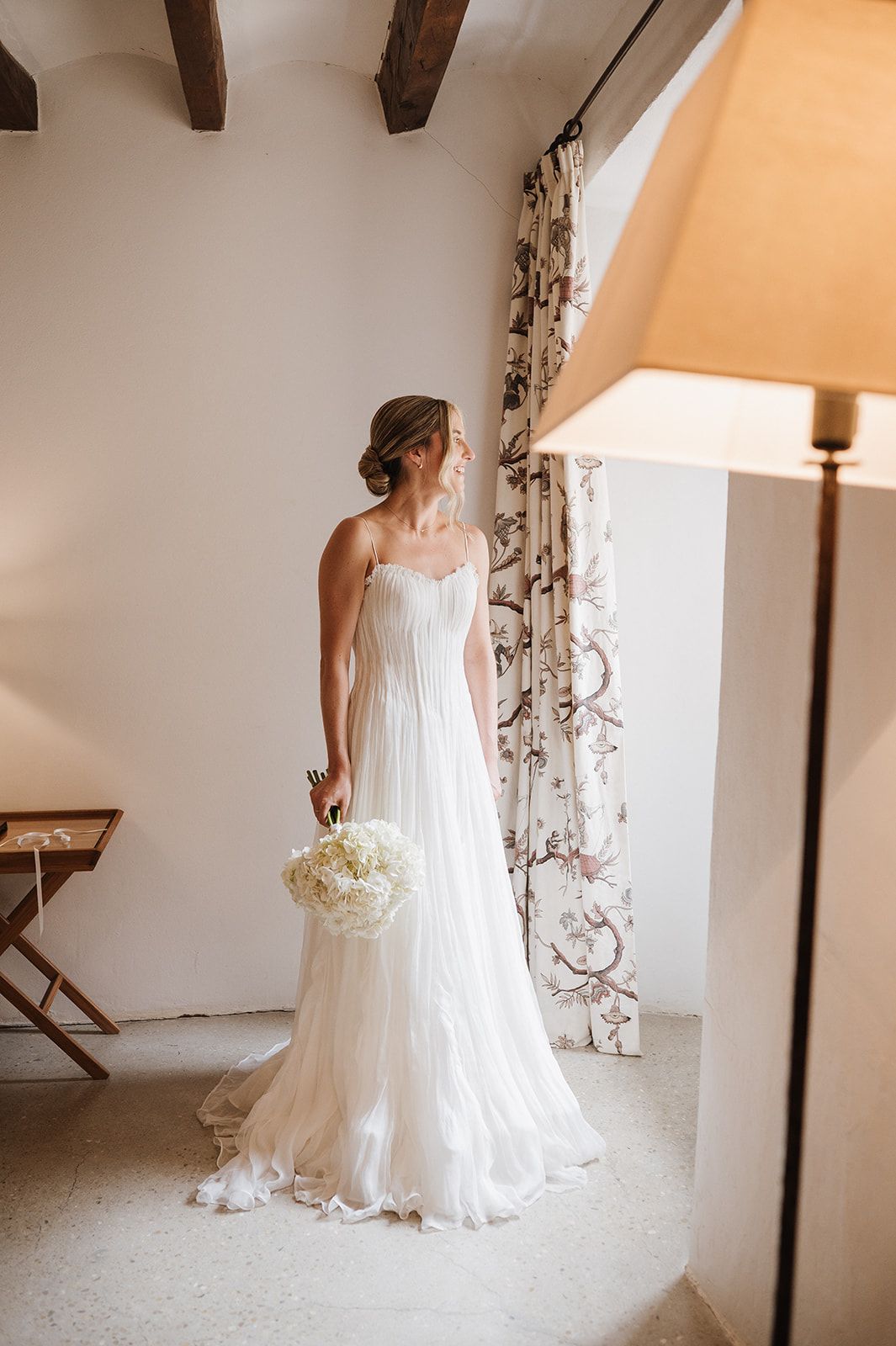 Bride in a flowing white strapless wedding gown holding a white hydrangea bouquet, standing in an elegant sunlit room.