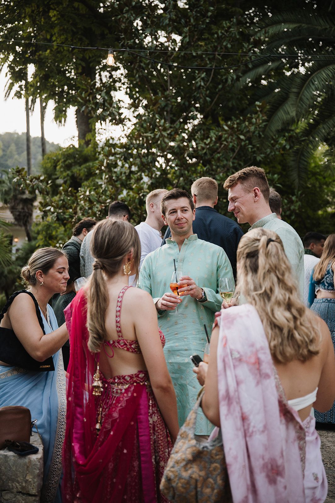 Wedding guests mingling outdoors in colorful Indian and western attire at garden reception.