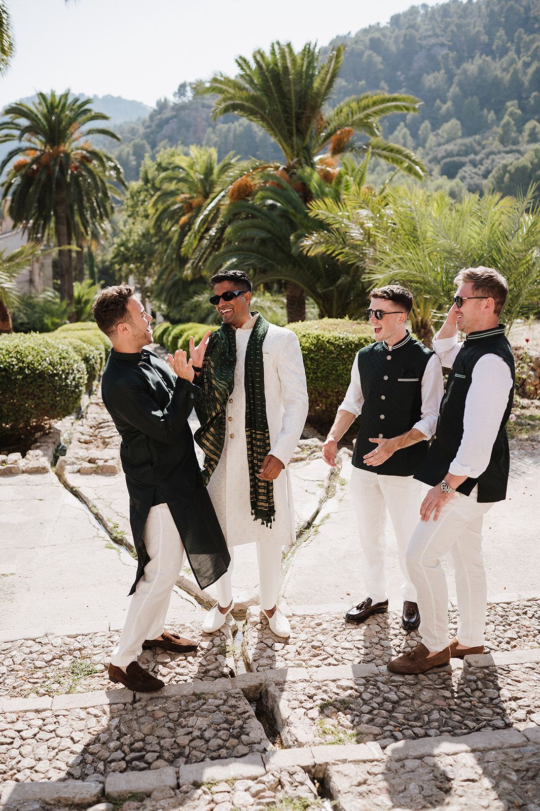 Groom and groomsmen in Indian wedding attire laughing outdoors among palm trees and lush greenery.