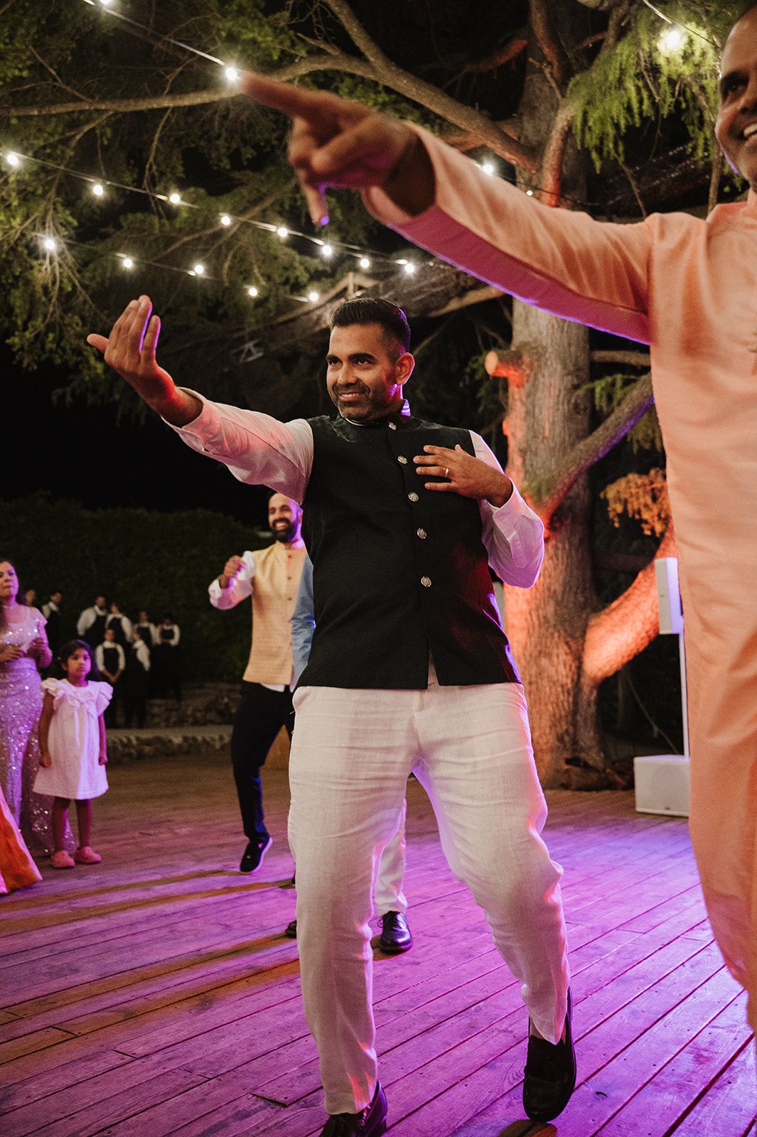 Man dancing joyfully in white kurta and navy blue nehru jacket at an outdoor Indian wedding reception.