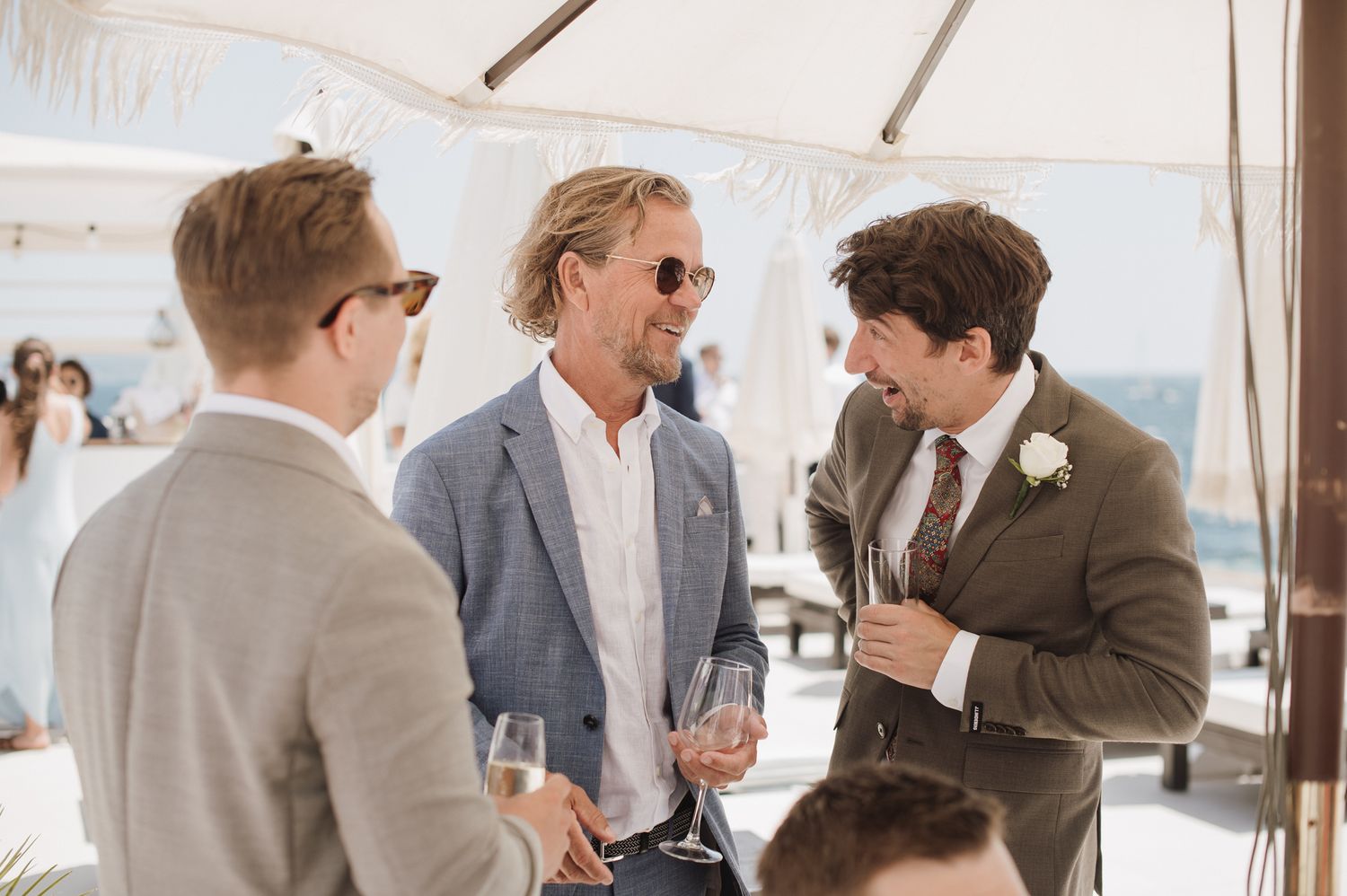 Wedding guests in suits chat and hold champagne glasses at sunny outdoor cocktail reception.
